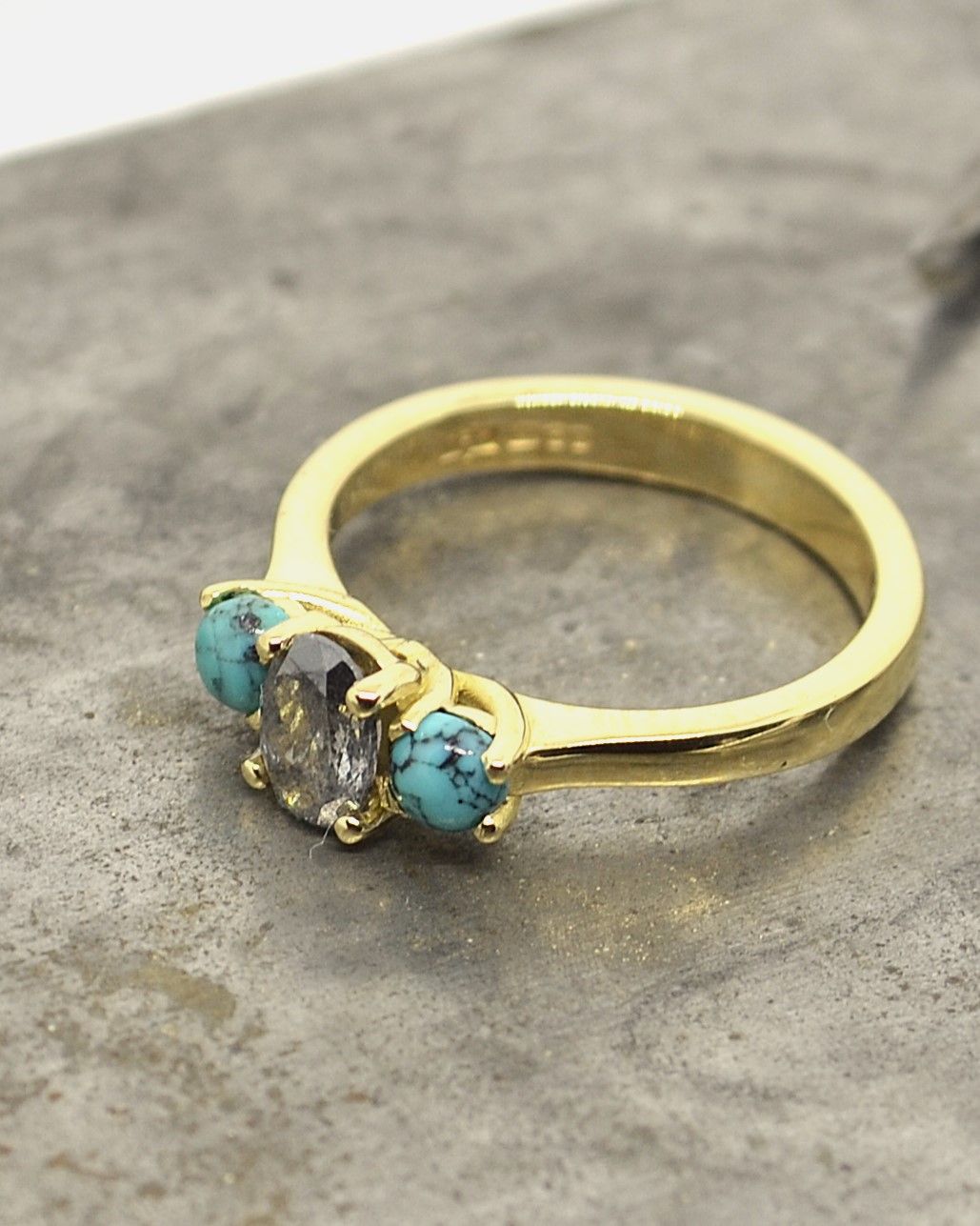 Handmade yellow gold ring featuring a rustic salt and pepper diamond with turquoise side stones, photographed on a textured metal surface