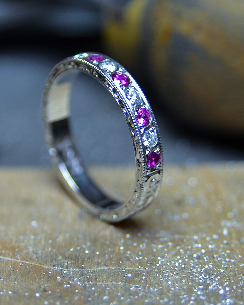 Platinum eternity ring with alternating round pink sapphires and white diamonds in a milgrain setting, photographed on a jeweller's workbench with a softly blurred workshop background.