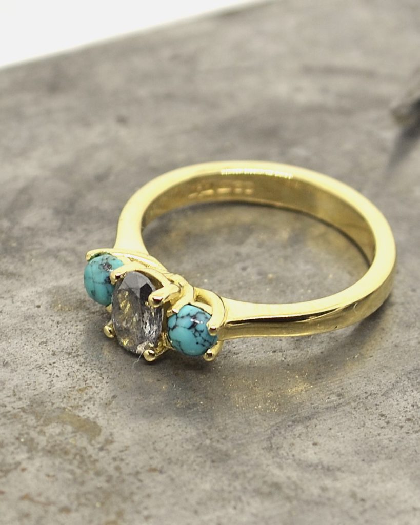 Handmade yellow gold ring featuring a rustic salt and pepper diamond with turquoise side stones, photographed on a textured metal surface